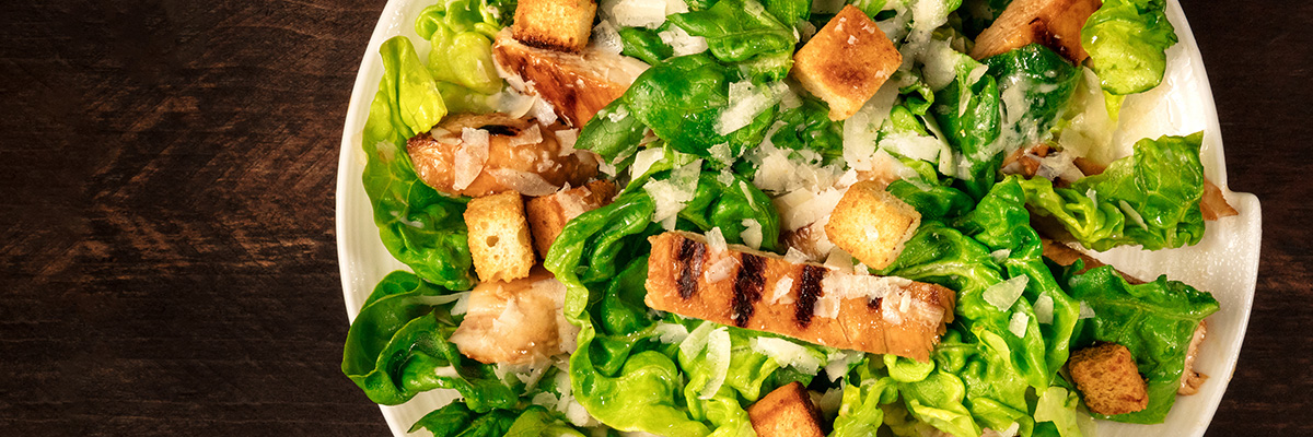 Kana-Caesarsalaatti krutongeilla ja parmesaanilla valkoisella lautasella rustiikkista taustaa vasten kuvattuna pystysuoraan ylhäältä